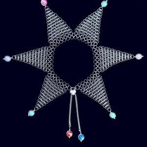 Puede incluir: Un collar de cota de malla plateado con un dise&ntilde;o de estrella. El collar presenta seis puntas triangulares, cada una adornada con una peque&ntilde;a cuenta iridiscente. Dos hebras con colgantes de coraz&oacute;n y cuentas cuelgan del centro. El fondo es azul marino oscuro.