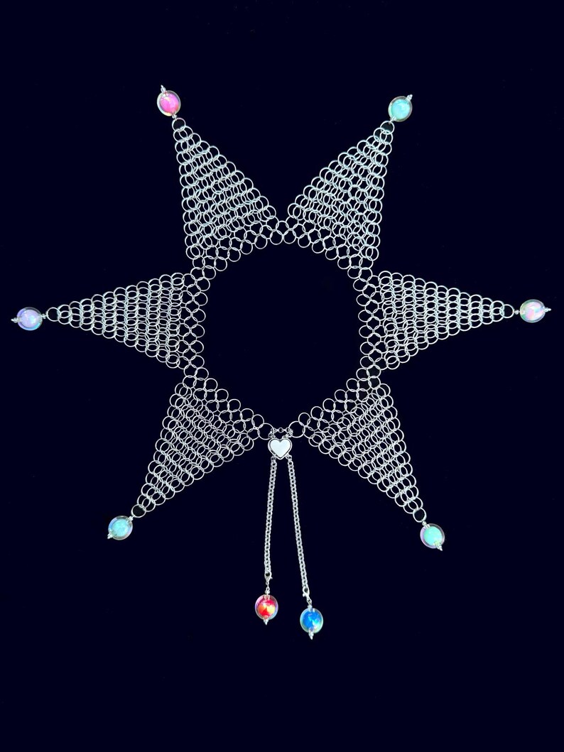 Puede incluir: Un collar de cota de malla plateado con un dise&ntilde;o de estrella. El collar presenta seis puntas triangulares, cada una adornada con una peque&ntilde;a cuenta iridiscente. Dos hebras con colgantes de coraz&oacute;n y cuentas cuelgan del centro. El fondo es azul marino oscuro.