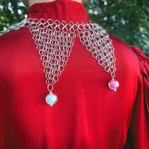 Puede incluir: Un collar de cota de malla plateada con dos colgantes de orbes de vidrio iridiscente. El collar se lleva sobre una blusa de sat&eacute;n roja. El fondo es un follaje verde borroso.