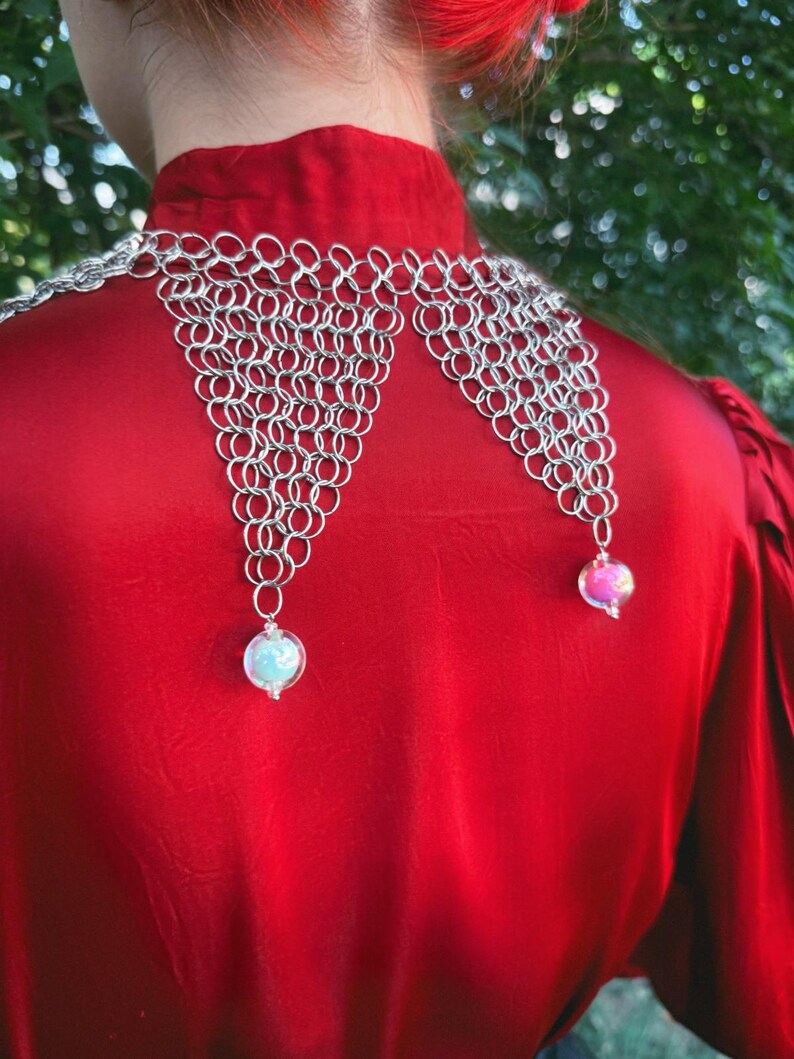Puede incluir: Un collar de cota de malla plateada con dos colgantes de orbes de vidrio iridiscente. El collar se lleva sobre una blusa de sat&eacute;n roja. El fondo es un follaje verde borroso.