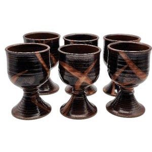 May include: Six brown ceramic goblets with a black and copper glaze. The goblets have a wide bowl and a short stem.