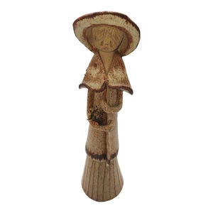 May include: A ceramic figurine of a woman wearing a brown and white dress and hat. She is holding a bouquet of flowers in her hand.