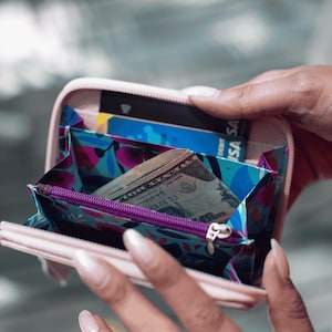 May include: A pink wallet with a floral pattern and a purple zipper is open, revealing a dollar bill and a blue credit card with the word "DEBIT VISA" printed on it.