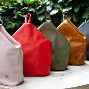 Puede incluir: Cinco mochilas de cuero en diferentes colores: rosa, rojo, verde, marrón y negro. Las mochilas tienen una forma triangular única y un pequeño logotipo en la parte delantera.