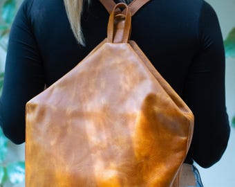 May include: A brown leather backpack with adjustable straps. The bag has a unique geometric shape and a small circular emblem on the front. The backpack is worn by a person wearing a black top and blue jeans.