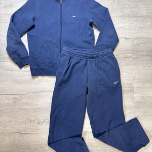K&ouml;nnte beinhalten: Ein marineblaues Nike-Trainingsanzug-Set. Die Jacke hat einen durchgehenden Rei&szlig;verschluss, Seitentaschen und ein kleines Nike-Logo auf der Brust. Die passende Hose hat einen elastischen Bund und ein Nike-Logo. Das Set ist aus weichem, bequemem Material.