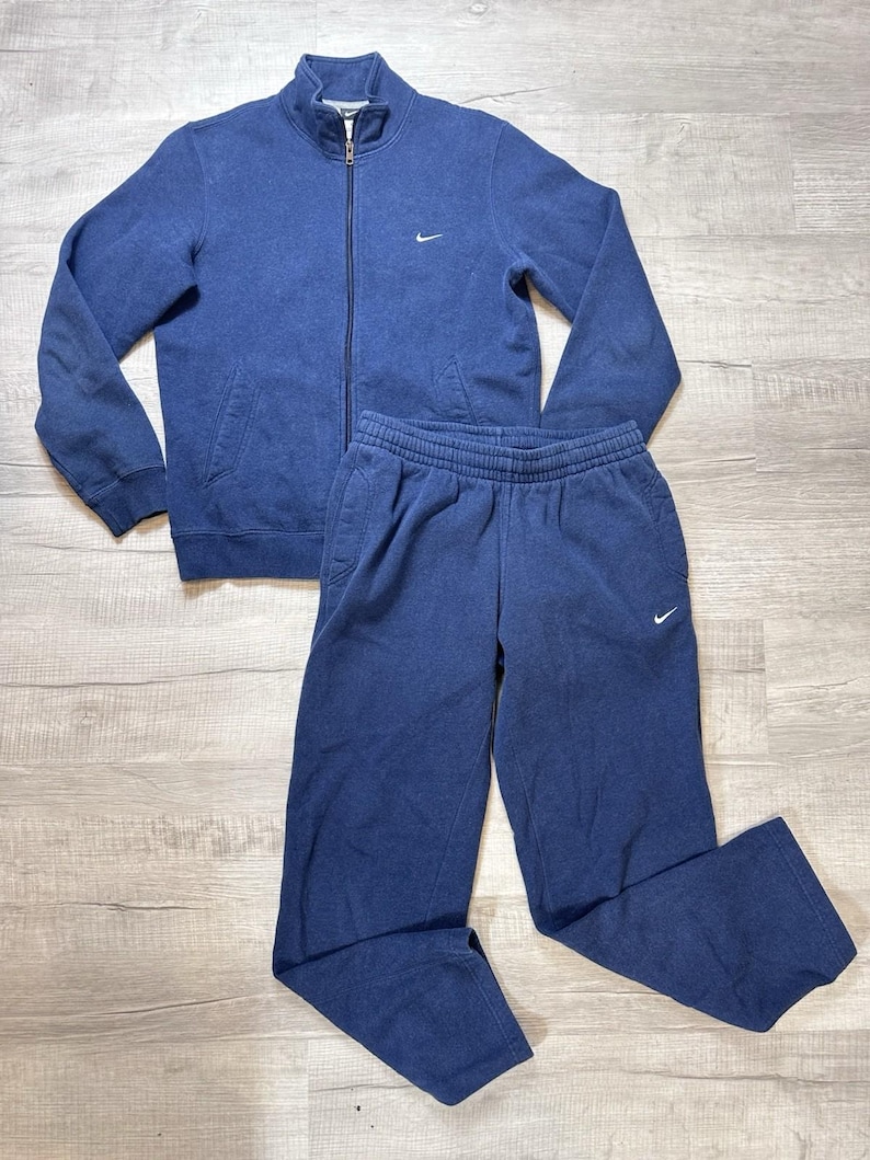 K&ouml;nnte beinhalten: Ein marineblaues Nike-Trainingsanzug-Set. Die Jacke hat einen durchgehenden Rei&szlig;verschluss, Seitentaschen und ein kleines Nike-Logo auf der Brust. Die passende Hose hat einen elastischen Bund und ein Nike-Logo. Das Set ist aus weichem, bequemem Material.