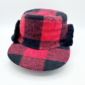 Pode incluir: Um boné de inverno com padrão xadrez vermelho e preto, aba reta e abas de orelha em pele sintética preta nas laterais. A copa e a aba são feitas de um tecido texturizado semelhante à lã, proporcionando uma estrutura bem definida.