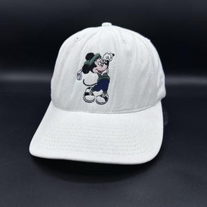May include: A white baseball cap featuring an embroidered graphic of a cartoon mouse wearing a green hat, a green sweater, and blue pants while holding a golf club. The cap has a curved brim and stitched detailing across the front panel.