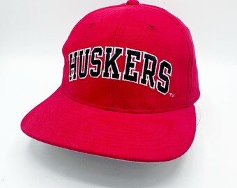 May include: A red baseball cap with the word "HUSKERS" in white block letters with a black outline. The hat has a curved brim and a button on top. The hat is a solid red color.