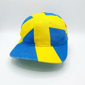 以下が含まれることがあります： 青と黄色の幾何学模様のベースボールキャップ。キャップはカーブしたつばと6パネル構造が特徴です。色は明るく、デザインは目を引きます。