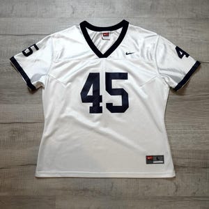 May include: White football jersey with navy blue accents, featuring the number 45 on the front and sleeves. The jersey has a V-neck collar and the Nike logo. The jersey is a size L.