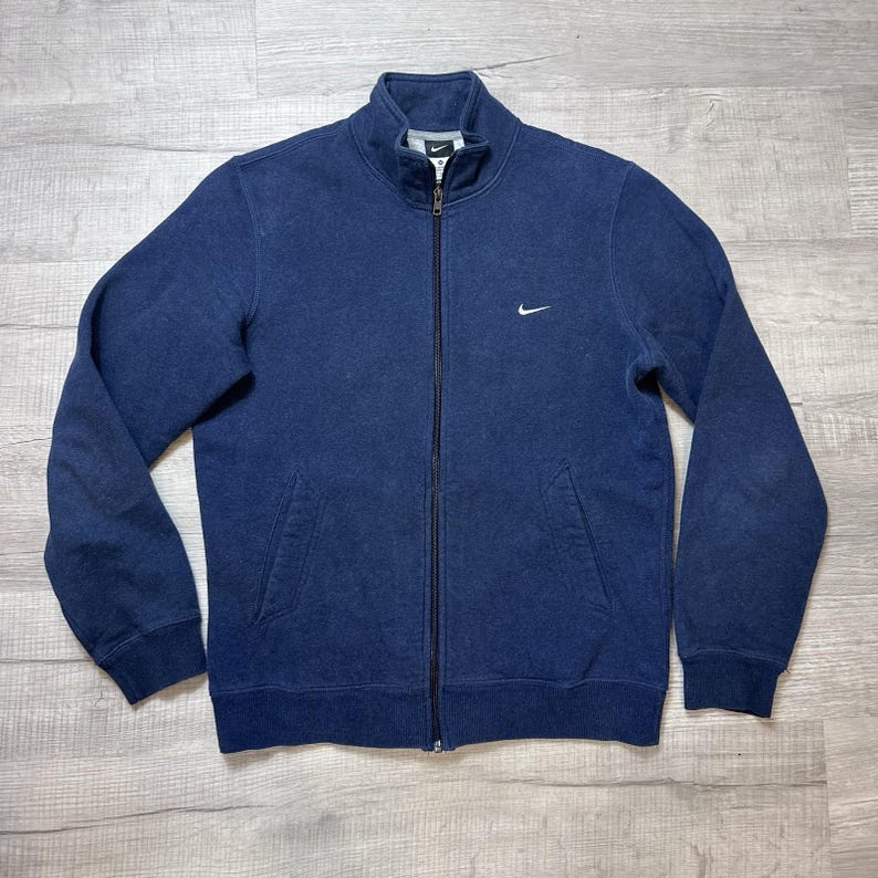 K&ouml;nnte beinhalten: Marineblaue Jacke mit Rei&szlig;verschluss, hohem Kragen und zwei Vordertaschen. Die Jacke hat ein kleines wei&szlig;es Nike-Logo auf der Brust. Sie ist aus einem weichen, strukturierten Stoff gefertigt.