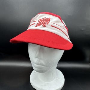 May include: A red and white visor with a red brim. The visor features a red letter "N" with a graphic on the front. Two red stripes are on the white portion of the visor.