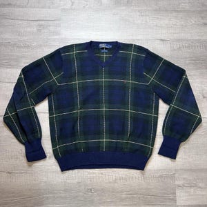 May include: A navy blue, green, and white plaid V-neck sweater. The sweater has long sleeves with navy blue cuffs and a navy blue hem. A small red logo is visible on the chest. The sweater is made of a soft, knitted material.