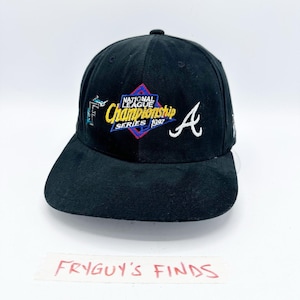 May include: A black baseball cap featuring an embroidered National League Championship Series 1997 logo in blue, yellow, and red. The cap includes a white Florida Marlins F logo on the left side and a white Atlanta Braves A logo on the right side.