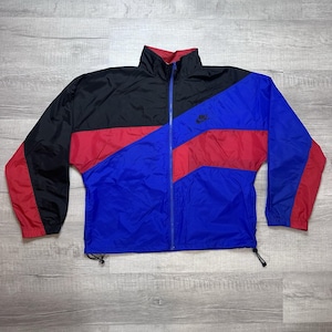 Op de afbeelding: Een vintage Nike windjack in zwart, rood en blauw. De jas heeft een volledige ritssluiting, een hoge kraag en het Nike-logo op de borst. Het colorblock-ontwerp omvat zwart aan de bovenkant, rood in het midden en blauw aan de onderkant.