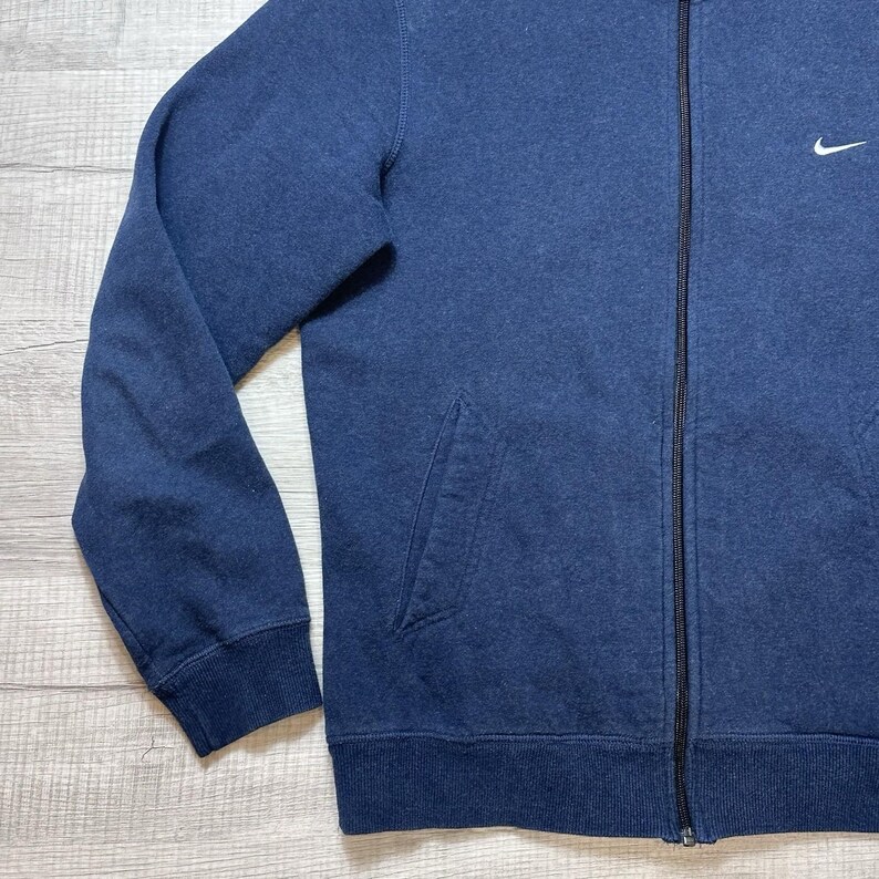 K&ouml;nnte beinhalten: Ein marineblauer Sweatshirt-Rei&szlig;verschluss mit einem kleinen wei&szlig;en Nike-Logo auf der oberen Brust. Das Sweatshirt hat einen durchgehenden Rei&szlig;verschluss, eine Vordertasche und gerippte B&uuml;ndchen und Saum. Der Stoff scheint weich und bequem zu sein.