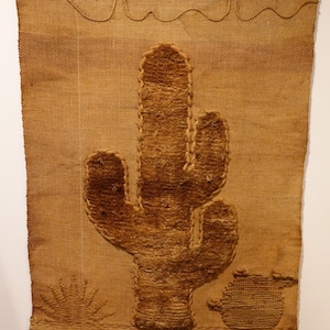 May include: A woven wall hanging featuring a large cactus design in shades of brown. The cactus is the focal point, with a sun and a small pot also depicted. The bottom edge has a fringe detail, and the top has a decorative border.