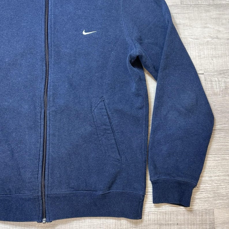 K&ouml;nnte beinhalten: Marineblaue Sweatshirt-Jacke mit Rei&szlig;verschluss und kleinem wei&szlig;en Nike-Logo auf der Brust. Mit Fronttasche und gerippten B&uuml;ndchen und Saum. Der Rei&szlig;verschluss ist schwarz.