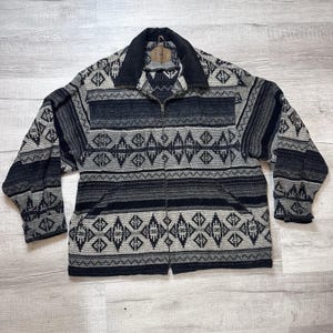 Puede incluir: Chaqueta con cremallera y estampado geométrico en tonos grises y negros. La chaqueta tiene cuello negro y cremallera completa. El diseño incluye motivos de diamantes y rayas, creando una apariencia texturizada. Fabricada con tejido grueso y tejido.