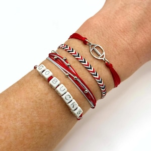 May include: A set of four bracelets featuring a red cord with a silver football charm, a braided red, white, and black cord, a red and white cord with silver beads, and a red cord with white square beads spelling out the text GO BUCKS.