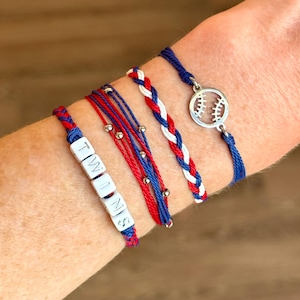 May include: A collection of five bracelets in red, white, and blue. One bracelet spells "TWINS" with white letter beads. Another features a baseball charm. Braided and beaded designs complete the set, perfect for sports fans.