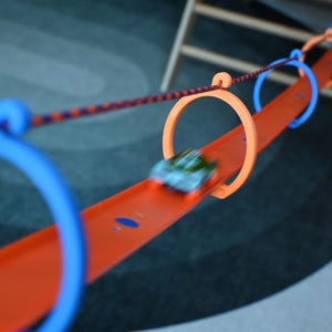 May include: An orange toy car track with blue and orange loops. A small, blurred toy car is moving along the track. A rope with purple and orange colors is attached to the track.