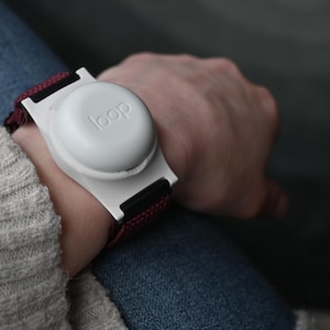 May include: A white wrist-worn device with the word "loop" printed on the top. The device is attached to a maroon and black strap. The device is worn on a person's wrist, with a gray sweater and blue jeans in the background.