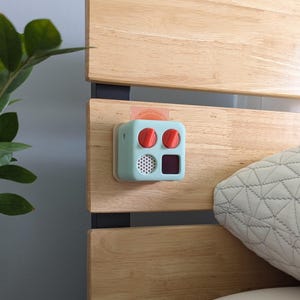 May include: A light blue and red sound machine with two red knobs and a small digital display. The sound machine is mounted on a wooden headboard.