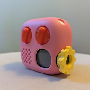 May include: A pink fidget cube featuring two orange toggle switches on the top, a small speaker grille on the left side, a dark circular screen in the center, and a yellow gear-shaped dial attached to the right side of the device.
