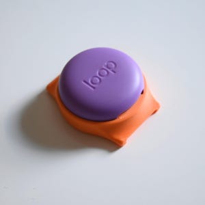 May include: A purple, circular object with the word "loop" embossed on top, resting in an orange, square-shaped holder. The holder has rounded edges and a textured appearance. The background is plain white.