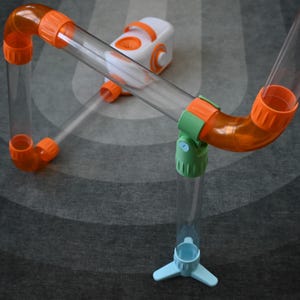 May include: A clear plastic toy with orange and green connectors and a white and orange base. The toy features clear tubes and curved orange pieces, connected by green and blue components. The toy is designed for children.