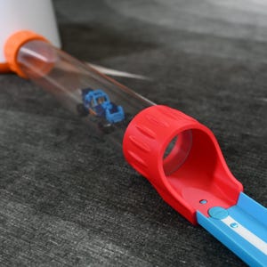 Puede incluir: Una pista de coches de juguete con una pista azul y blanca, un bucle rojo y un tubo de plástico transparente. Un pequeño camión de juguete azul está dentro del tubo. La pista está sobre una superficie gris.
