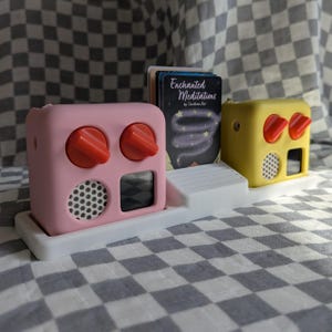 May include: Two pink and yellow toy radios with red knobs and a white base. The radios are on a black and white chequered surface. The pink radio has a black screen and a white speaker grille. The yellow radio has a black screen and a white speaker grille. The radios are labelled "Enchanted Meditations by Christine Kane".