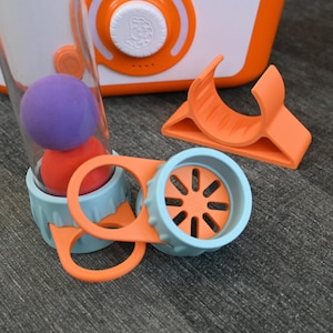 Peut inclure: Un jouet orange et blanc avec un cylindre transparent contenant une boule violette et rouge. Des composants orange et bleus supplémentaires sont dispersés autour du jouet. Le jouet a un cadran et des boutons. Le fond est une surface grise.