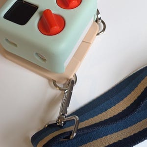May include: A light blue camera case with a beige strap and two red buttons. The case has a small screen and a silver clip.