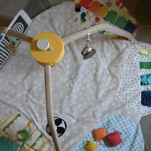 May include: A baby play gym with a yellow mobile and a soft, patterned play mat. The mobile has wooden arms and hanging toys. The mat features various textures and colorful patches, including a black and white pattern and a soccer ball graphic.