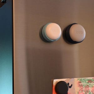 May include: Two circular magnetic buttons attached to a metallic surface. One button has a light blue base and a white top, while the other has a dark blue base and a white top. Both white tops feature the word loop embossed in lowercase letters.