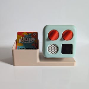 May include: A light blue toy with two red knobs, a speaker with a black mesh grille, and a black screen. The toy is sitting on a beige stand with a card that says "Ladybird Audio Adventures Rainforests" in it.