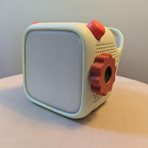 May include: A pale yellow, cube-shaped electronic device with a large, smooth, light-colored front panel. It features a prominent orange gear-shaped dial on the side, two orange rounded buttons on top, and a perforated speaker grille.