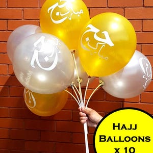May include: A hand holds a bunch of ten balloons in gold and silver. The gold balloons have white Arabic script. The balloons are against a brick wall. The text "HAJJ BALLOONS x 10" is in a yellow circle.