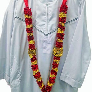 May include: A white long-sleeved garment with a central zip and a decorative V-shaped trim, draped with a long garland featuring alternating red and gold fabric ruffles and rosettes arranged in a continuous, repeating pattern.