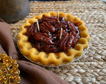 May include: A pecan pie candle in a yellow pie crust with a brown pecan filling and three white wicks.