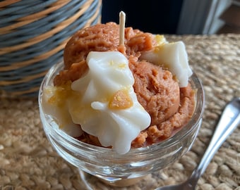 May include: A glass dessert dish filled with a brown and white candle shaped like ice cream with whipped cream and caramel sauce.