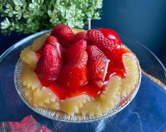 May include: A strawberry shortcake candle with red frosting and white candles. The candle is in a silver pie pan and is on a glass cake stand.