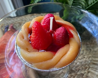 May include: A strawberry shortcake candle in a silver foil pie pan. The candle is decorated with three red strawberries and a white wick.
