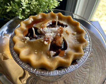 May include: A brown pie-shaped candle with a brown glaze and white sprinkles. The candle has three wicks and is sitting on a clear glass plate.