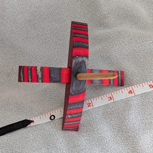 May include: A wooden toy top with a cross-shaped design. The top has alternating red and grey stripes. A wooden dowel is inserted through the centre. A measuring tape is next to the top.