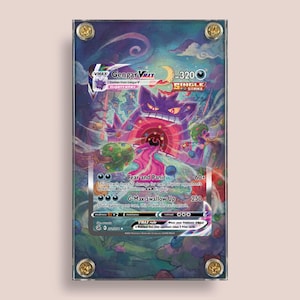 May include: A framed Pokémon card featuring Gengar VMAX. The card has a purple and pink color scheme with the text "Fear and Panic" and "G-Max Swallow Up". The card is encased in a clear acrylic frame with gold-colored hardware.